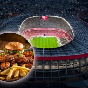 Composición Fotográfica Horizontal Que Muestra Una Vista Aérea Nocturna Del Estadio Akron En Guadalajara, Con Su Característica Estructura Iluminada Y La Cancha De Fútbol Verde Brillante Visible En El Centro. En La Esquina Inferior Izquierda Se Superpone Un Círculo Recortado Que Resalta Un Menú De Comida Rápida Sobre Una Superficie De Madera, Compuesto Por Alitas De Pollo Fritas, Una Porción Abundante De Papas A La Francesa Y Dos Hamburguesas Con Ajonjolí, Lechuga Y Jitomate Al Fondo.