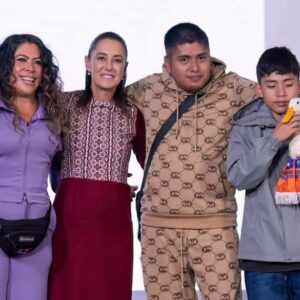 Una Fotografía Grupal De Cuatro Personas Y Un Pato Doméstico Posando Juntas En Un Escenario. De Izquierda A Derecha: Una Mujer Con Un Traje Deportivo Morado De Dos Piezas, Claudia Sheinbaum Con Un Vestido Estampado Rojo Y Blanco Con Una Falda Burdeos, Un Hombre Joven Con Un Traje De Chándal Estampado Marrón Y Beige, Y Un Joven Con Una Chaqueta Gris Sosteniendo Al Pato Doméstico Merlín, Que Lleva Un Chaleco Verde Y Una Bufanda Naranja. Se Paran Frente A Un Fondo Blanco Con Texto Parcial Y Una Bandera Mexicana.