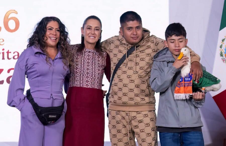Una Fotografía Grupal De Cuatro Personas Y Un Pato Doméstico Posando Juntas En Un Escenario. De Izquierda A Derecha: Una Mujer Con Un Traje Deportivo Morado De Dos Piezas, Claudia Sheinbaum Con Un Vestido Estampado Rojo Y Blanco Con Una Falda Burdeos, Un Hombre Joven Con Un Traje De Chándal Estampado Marrón Y Beige, Y Un Joven Con Una Chaqueta Gris Sosteniendo Al Pato Doméstico Merlín, Que Lleva Un Chaleco Verde Y Una Bufanda Naranja. Se Paran Frente A Un Fondo Blanco Con Texto Parcial Y Una Bandera Mexicana.