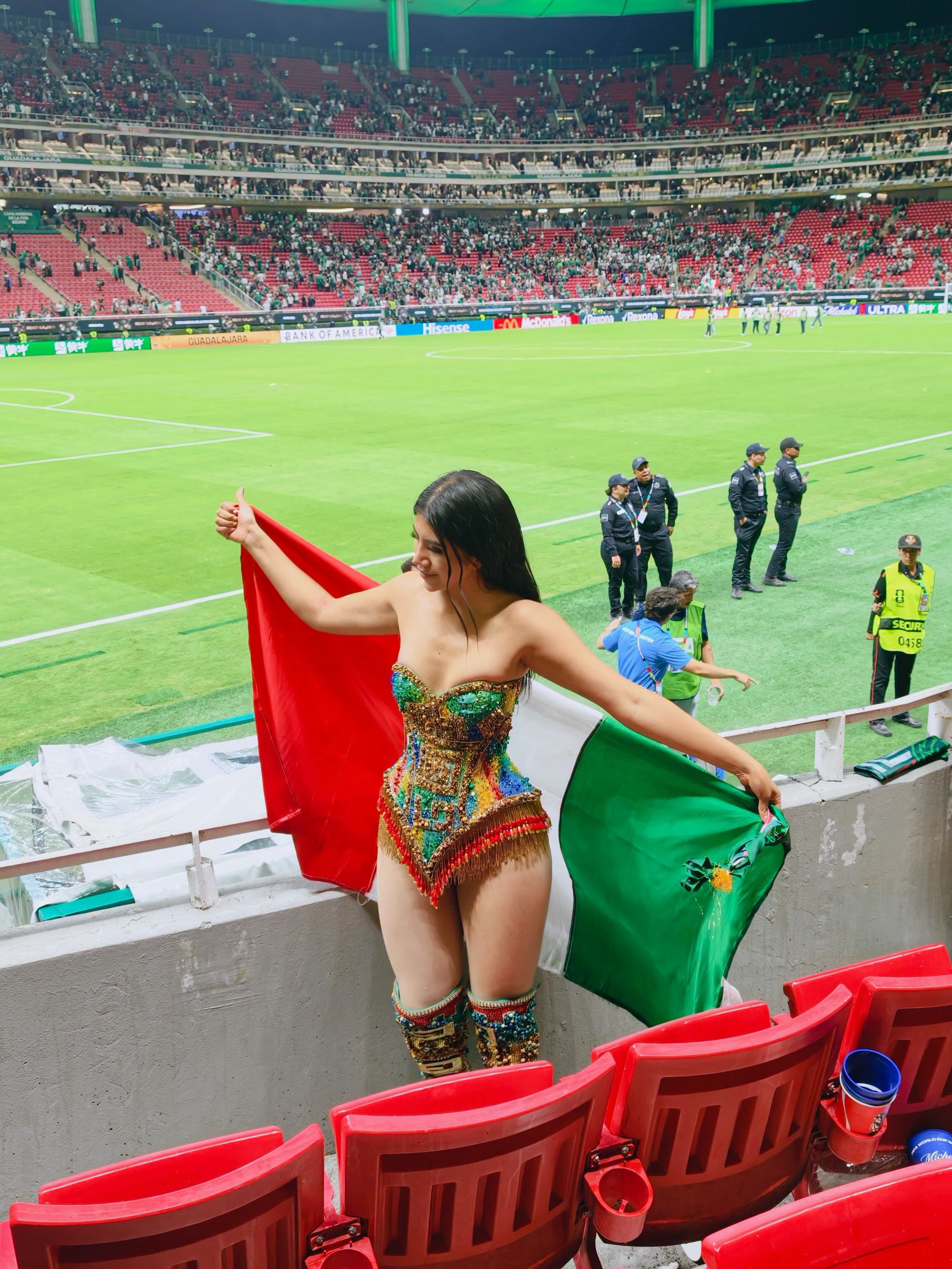¡de calkiní para el mundo! Diseño campechano brilla en el estadio akron durante el mundial 2026 la revista del sureste mx La Influencer Friday Frida Posa Desde Las Gradas Del Estadio Akron Sosteniendo Con Los Brazos Abiertos Una Bandera De México. Viste El Traje Artesanal De Alberto Cuevas, Un Body Tipo Corsé Strapless Multicolor Cubierto De Pedrería Y Flecos Dorados, Acompañado De Botas Altas A Juego Con El Mismo Diseño. Al Fondo Se Aprecia La Cancha De Fútbol Y Las Tribunas Del Estadio Llenas De Aficionados.
