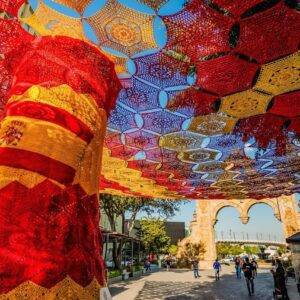 El arte que abraza al fútbol: zapopan inaugura un monumental cielo tejido por manos mexicanas para el mundial 2026 la revista del sureste mx Una Fotografía En Contrapicado Que Captura De Cerca Un Monumental &Amp;Quot;Cielo Tejido&Amp;Quot; Suspendido Entre Árboles Frondosos Bajo Un Cielo Despejado. La Instalación Artística Está Compuesta Por Miles De Carpetas O Hexágonos Tejidos A Mano En Técnica De Crochet Que Se Entrelazan Para Formar Un Gran Manto Calado. En El Centro De La Composición Destaca Una Sección Circular Tejida Con Los Colores Azul Y Rojo, Rodeada Por Extensiones Textiles En Color Blanco, Amarillo Y Verde. Las Hojas Verdes De Los Árboles Enmarcan Los Costados De La Colorida Estructura Artesanal.