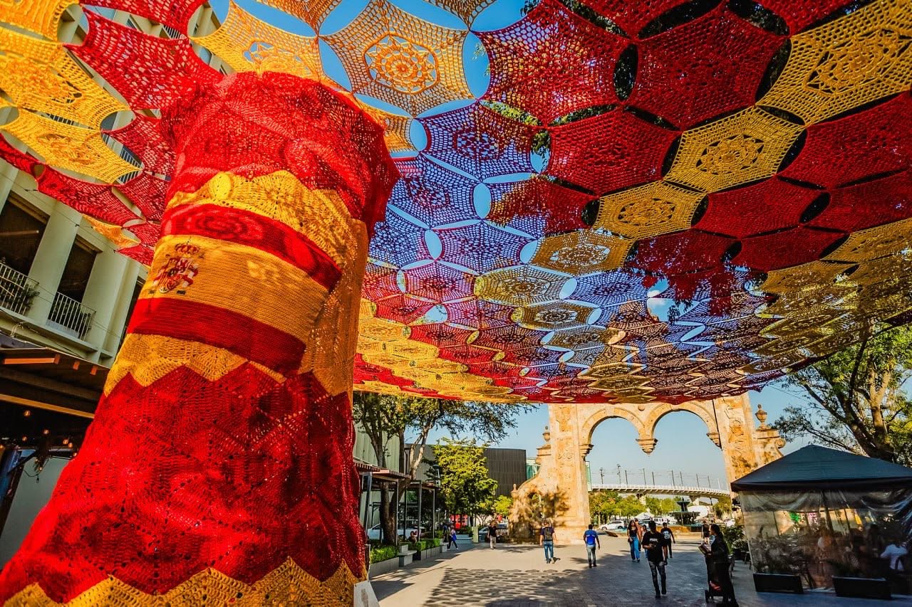 Una Fotografía En Contrapicado Que Captura De Cerca Un Monumental &Quot;Cielo Tejido&Quot; Suspendido Entre Árboles Frondosos Bajo Un Cielo Despejado. La Instalación Artística Está Compuesta Por Miles De Carpetas O Hexágonos Tejidos A Mano En Técnica De Crochet Que Se Entrelazan Para Formar Un Gran Manto Calado. En El Centro De La Composición Destaca Una Sección Circular Tejida Con Los Colores Azul Y Rojo, Rodeada Por Extensiones Textiles En Color Blanco, Amarillo Y Verde. Las Hojas Verdes De Los Árboles Enmarcan Los Costados De La Colorida Estructura Artesanal.
