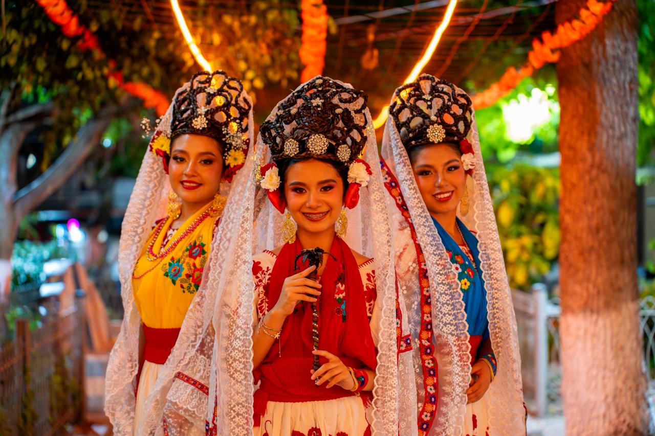 Fotografía De Plano Medio En Exteriores Que Muestra A Tres Mujeres Jóvenes Sonriendo, Vestidas Con El Traje Tradicional De La Huasteca Hidalguense. Están Alineadas De Frente A La Cámara; La Joven De La Izquierda Viste Una Blusa Amarilla Con Bordados Florales, La Del Centro Lleva Una Blusa Roja Y Sostiene Un Abanico Artesanal Oscuro, Y La De La Derecha Usa Una Blusa Azul Rey. Las Tres Portan Faldas Blancas Con Bordados, Collares Dorados, Aretes Largos Y Elaborados Tocados Prehispánicos Entrelazados En El Cabello Con Flores, Cubiertos Por Largos Velos De Encaje Blanco Que Caen Sobre Sus Hombros. El Fondo Está Sutilmente Desenfocado, Mostrando Un Entorno Festivo Nocturno Iluminado Por Una Serie De Luces Cálidas Arqueadas Y Decoraciones De Color Naranja.