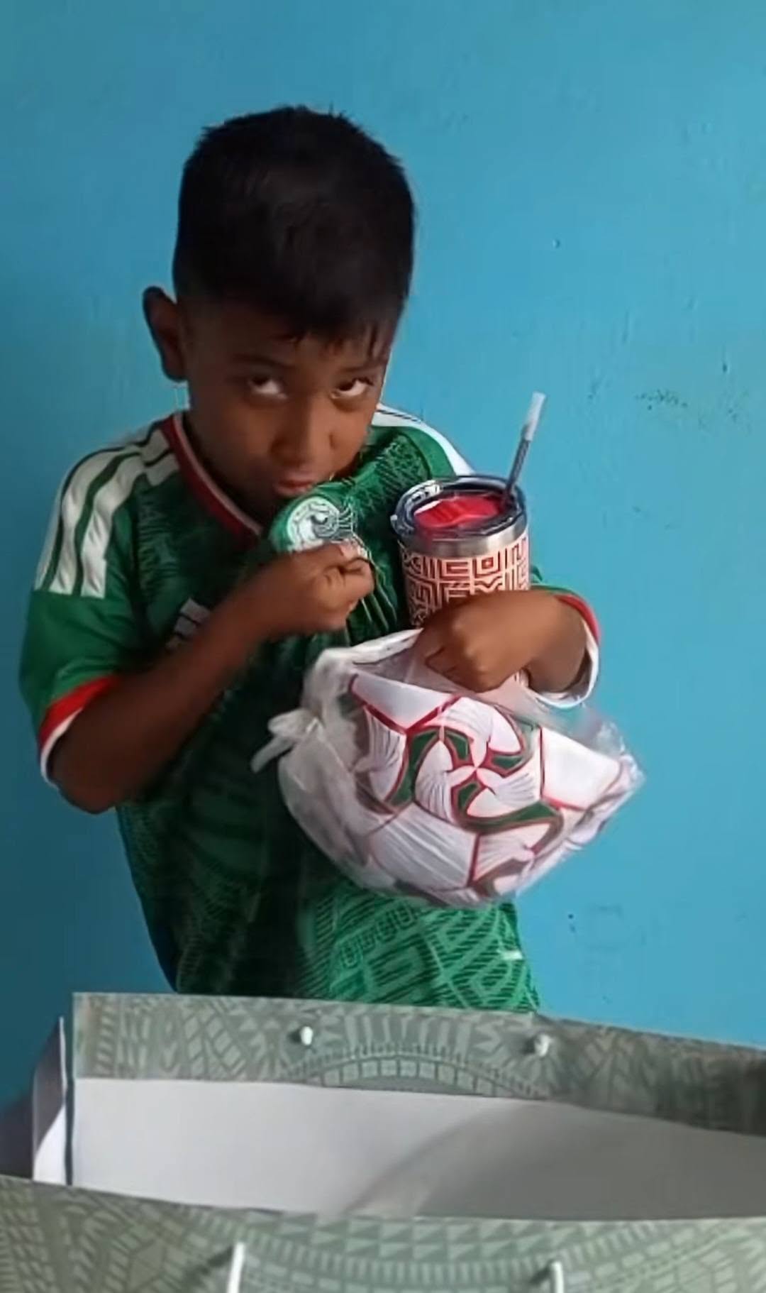 El regalo del tri al niño que hizo su playera con una bolsa de papas: iker conmueve al mundial 2026 la revista del sureste mx Un Niño Sonriente, Vistiendo La Playera Verde Oficial De La Selección Mexicana, Sostiene Emocionado Un Balón De Fútbol Dentro De Su Bolsa Transparente Y Un Termo Conmemorativo De Color Rojo Y Plata, Mientras Besa Con Ternura El Escudo Del Tri En Su Camiseta Frente A Una Pared Azul.