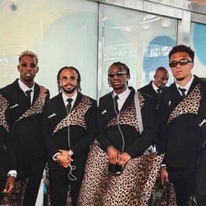 El regreso (con mucho estilo) de la república democrática del congo al mundial 2026 la revista del sureste mx Fotografía De Seis Integrantes De La Selección De Futbol De La República Democrática Del Congo Posando En Un Aeropuerto. Todos Visten Elegantes Trajes Sastre De Color Negro Con Una Banda Diagonal Estampada Con Patrón De Leopardo En El Pecho Y Un Broche Brillante. Llevan Gafas De Sol, Maletas De Viaje Metálicas Y Bolsos Grandes Con El Mismo Diseño De Leopardo.