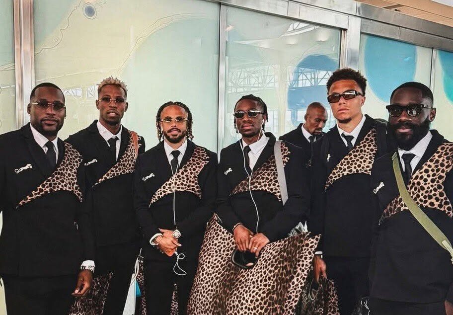 El regreso (con mucho estilo) de la república democrática del congo al mundial 2026 la revista del sureste mx Fotografía De Seis Integrantes De La Selección De Futbol De La República Democrática Del Congo Posando En Un Aeropuerto. Todos Visten Elegantes Trajes Sastre De Color Negro Con Una Banda Diagonal Estampada Con Patrón De Leopardo En El Pecho Y Un Broche Brillante. Llevan Gafas De Sol, Maletas De Viaje Metálicas Y Bolsos Grandes Con El Mismo Diseño De Leopardo.