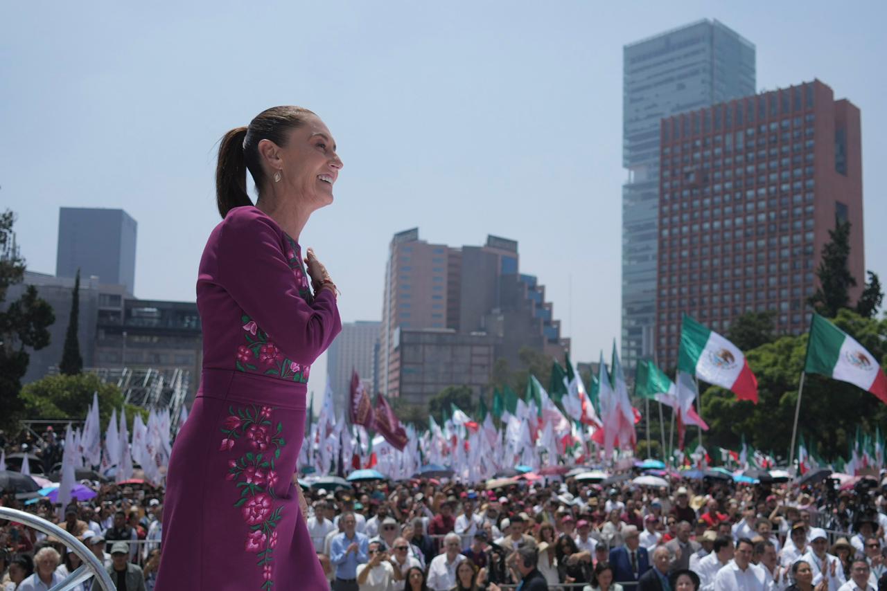 Hilos de identidad: el significado detrás del vestido oaxaqueño de la presidenta claudia sheinbaum la revista del sureste mx Una Toma De Cuerpo Entero Y Perfil De La Presidenta De México, Claudia Sheinbaum, En Un Mitin Al Aire Libre. Ella Está De Pie Sobre Una Plataforma Elevada, Mirando Hacia La Derecha Y Sonriendo Con La Mano Derecha Sobre El Corazón, De Espaldas A Una Multitud Densa Y Ondeando Banderas (Incluidas Banderas Mexicanas Y Blancas). Viste Un Vestido De Manga Larga De Color Fucsia Brillante Con Intrincados Bordados Florales En Tonos Fucsia Y Verde En El Corpiño Y La Falda.