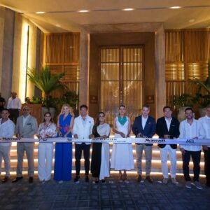 Fotografía De Plano Entero En Formato Horizontal Que Muestra El Corte De Listón Inaugural Por La Noche. Un Grupo De Catorce Autoridades Y Empresarios De La Industria Turística Están Alineados De Pie Frente Al Acceso Principal De Un Hotel De Ultralujo. El Fondo Destaca Por Una Imponente Fachada Arquitectónica Iluminada Con Luces Cálidas, Techos Altos, Columnas De Piedra Y Grandes Puertas De Madera Con Rejillas Verticales, Flanqueadas Por Plantas Tropicales. En El Centro, La Gobernadora Mara Lezama Sonríe Sosteniendo Las Tijeras Junto A Una Cinta Blanca Que Cruza De Extremo A Extremo Con Los Logotipos De St. Regis Y Marriott International. Los Asistentes Visten Atuendos Formales, Trajes Ligeros En Tonos Claros Y Vestidos Elegantes Ideales Para El Clima Del Caribe Mexicano.
