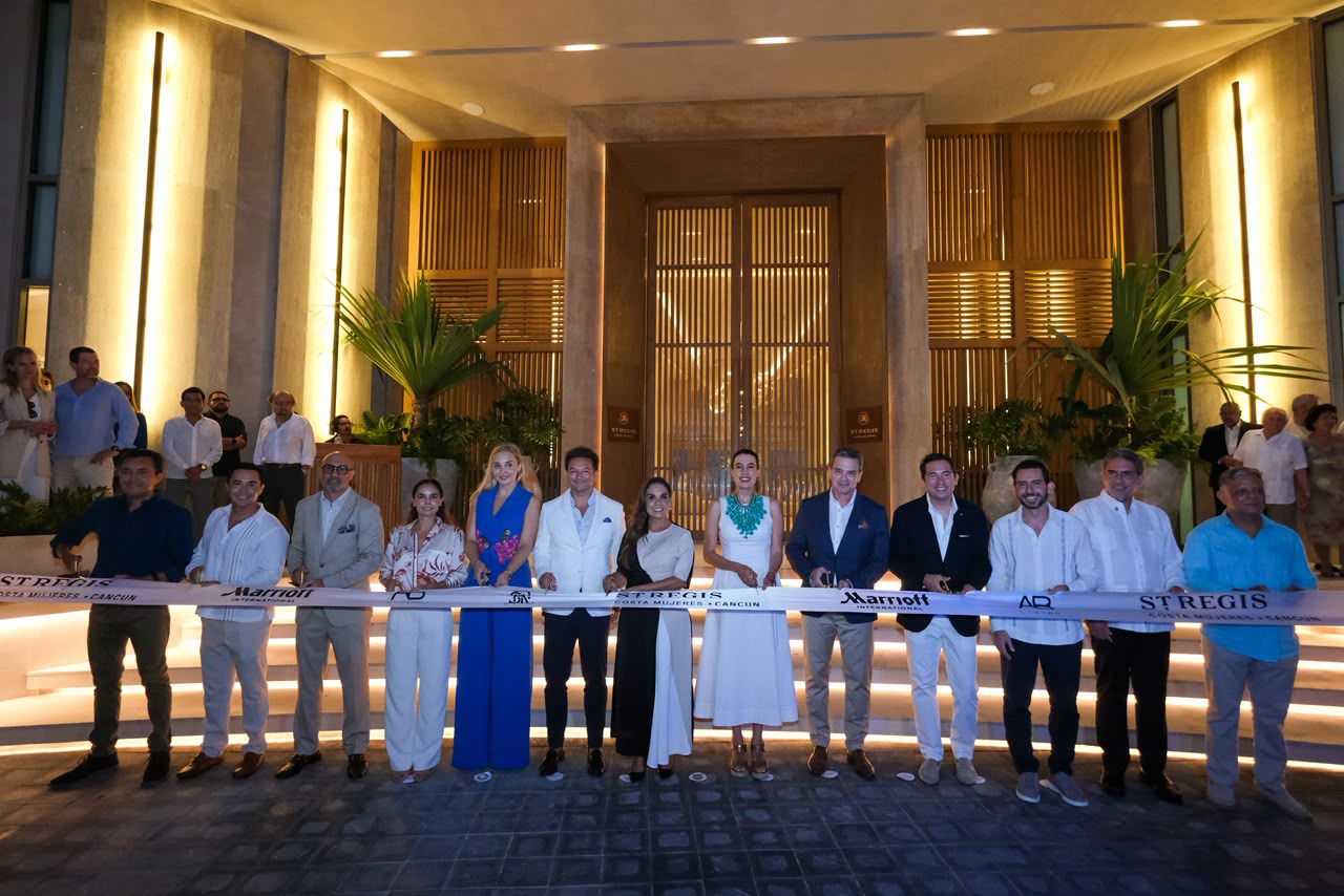 Fotografía De Plano Entero En Formato Horizontal Que Muestra El Corte De Listón Inaugural Por La Noche. Un Grupo De Catorce Autoridades Y Empresarios De La Industria Turística Están Alineados De Pie Frente Al Acceso Principal De Un Hotel De Ultralujo. El Fondo Destaca Por Una Imponente Fachada Arquitectónica Iluminada Con Luces Cálidas, Techos Altos, Columnas De Piedra Y Grandes Puertas De Madera Con Rejillas Verticales, Flanqueadas Por Plantas Tropicales. En El Centro, La Gobernadora Mara Lezama Sonríe Sosteniendo Las Tijeras Junto A Una Cinta Blanca Que Cruza De Extremo A Extremo Con Los Logotipos De St. Regis Y Marriott International. Los Asistentes Visten Atuendos Formales, Trajes Ligeros En Tonos Claros Y Vestidos Elegantes Ideales Para El Clima Del Caribe Mexicano.