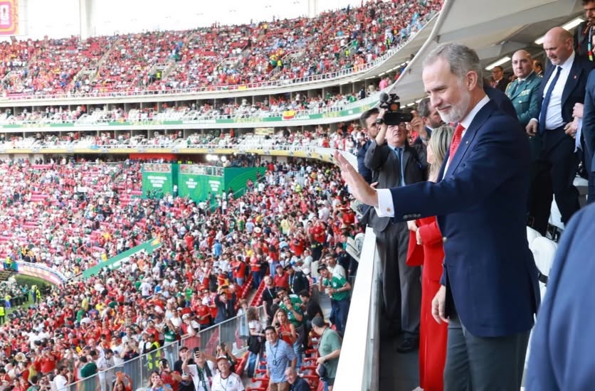 Fotografía En Plano Medio Largo Que Muestra De Perfil Al Rey Felipe Vi De España En Un Palco Del Estadio Guadalajara Durante Un Partido Del Mundial 2026. El Monarca, Vestido Con Un Traje Sastre Azul Marino, Camisa Blanca Y Corbata Roja, Sonríe Y Saluda Con La Mano Derecha Extendida Hacia La Multitud. Detrás De Él Se Encuentran Otros Asistentes Con Trajes Formales Y Personal De Seguridad. A La Izquierda, El Fondo Muestra Las Gradas Del Estadio Completamente Llenas De Aficionados Vistiendo Camisetas En Su Mayoría Rojas, Verdes Y Blancas Bajo Una Iluminación Diurna De Exterior.