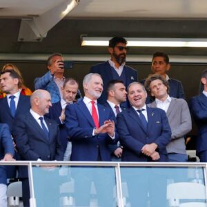 Fotografía De Plano Medio Largo En Formato Horizontal Que Muestra El Interior De Un Palco Techado En El Estadio Guadalajara. En El Centro, En Primera Fila, Aparece El Rey Felipe Vi De España Sonriendo Y Aplaudiendo, Vestido Con Traje Azul Marino, Camisa Blanca Y Corbata Roja. A Su Izquierda Y Derecha Hay Un Nutrido Grupo De Invitados Especiales Y Autoridades Con Trajes Formales, Entre Ellos Varios Exjugadores De Fútbol, Quienes Conversan Y Sonríen. En La Fila Posterior, Se Observa A Un Hombre Con Barba Y Lentes De Sol Y A Otros Asistentes Observando El Terreno De Juego. Una Barandilla De Vidrio Delimita La Parte Frontal Del Palco.