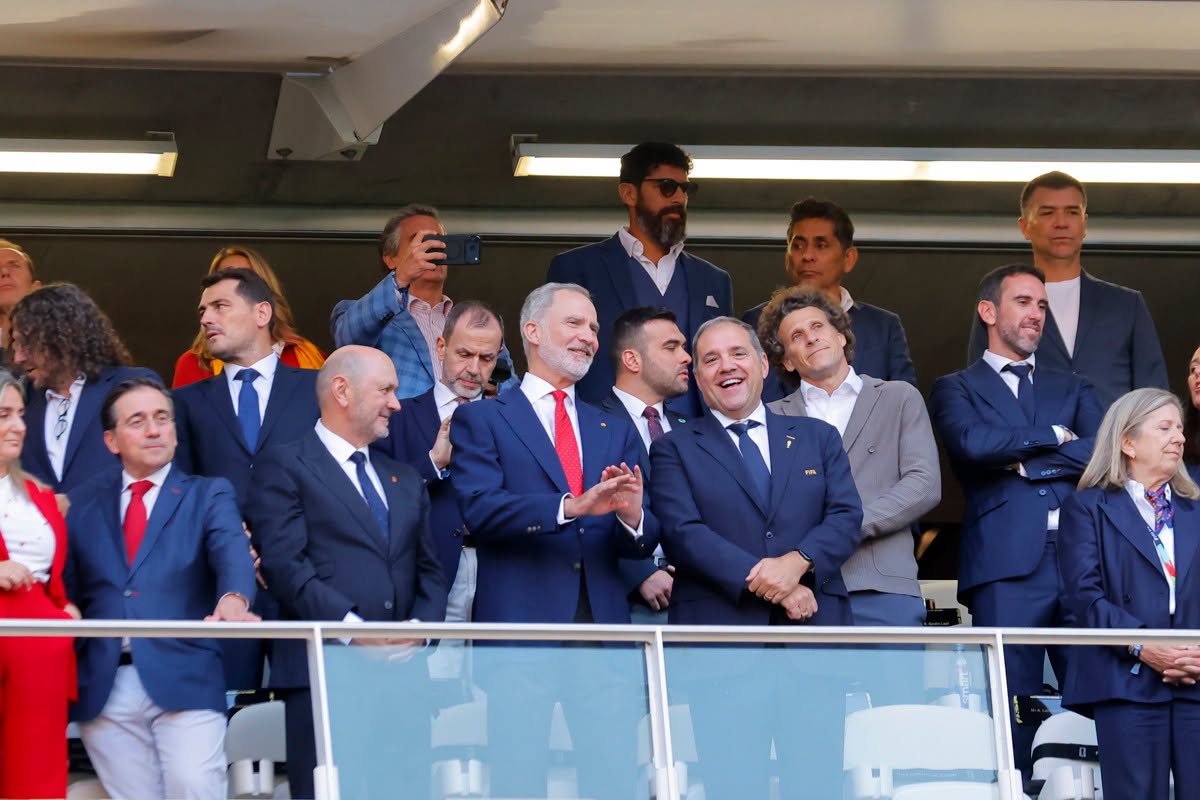 Fotografía De Plano Medio Largo En Formato Horizontal Que Muestra El Interior De Un Palco Techado En El Estadio Guadalajara. En El Centro, En Primera Fila, Aparece El Rey Felipe Vi De España Sonriendo Y Aplaudiendo, Vestido Con Traje Azul Marino, Camisa Blanca Y Corbata Roja. A Su Izquierda Y Derecha Hay Un Nutrido Grupo De Invitados Especiales Y Autoridades Con Trajes Formales, Entre Ellos Varios Exjugadores De Fútbol, Quienes Conversan Y Sonríen. En La Fila Posterior, Se Observa A Un Hombre Con Barba Y Lentes De Sol Y A Otros Asistentes Observando El Terreno De Juego. Una Barandilla De Vidrio Delimita La Parte Frontal Del Palco.