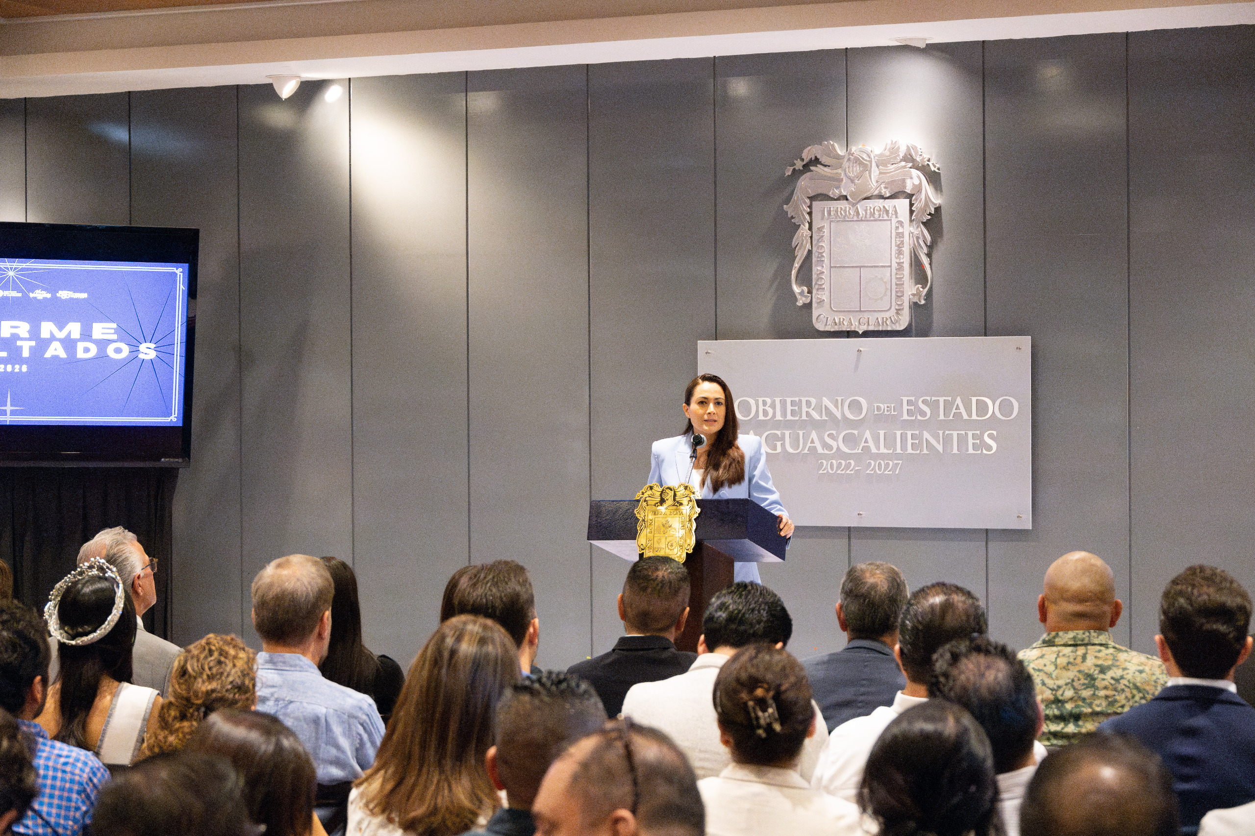 Feria nacional de san marcos 2026: una edición histórica que rompe récords y conquista al mundo la revista del sureste mx Una Fotografía Tomada Desde La Perspectiva Del Público En Un Auditorio. Al Fondo, La Gobernadora Tere Jiménez, Vistiendo Un Saco Azul Claro, Se Encuentra De Pie Detrás De Un Podio De Madera Oscura Con Un Escudo Dorado, Hablando Frente A Un Micrófono. Detrás De Ella Hay Un Panel Gris Con Letras Blancas Que Dicen &Quot;Gobierno Del Estado Aguascalientes 2022 - 2027&Quot; Bajo Un Escudo De Armas Plateado.
