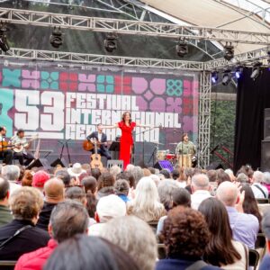 Una Toma De Plano General Desde La Perspectiva De La Audiencia Muestra Un Concierto De Música En Vivo En Un Escenario Al Aire Libre Cubierto Por Una Carpa Translúcida. En El Escenario, Una Cantante Con Un Vestido Largo De Color Rojo Brillante Se Encuentra En El Centro Con Los Brazos Abiertos, Actuando Frente A Un Micrófono.