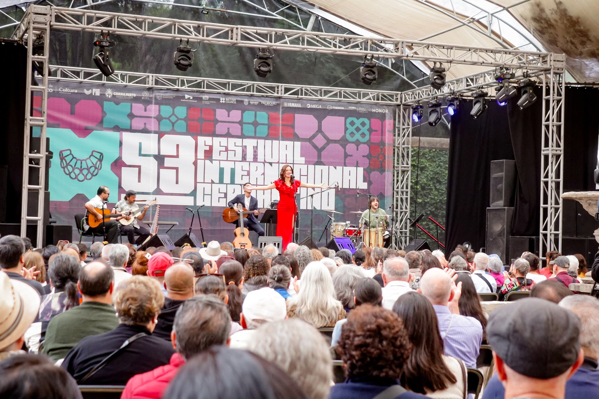 Una Toma De Plano General Desde La Perspectiva De La Audiencia Muestra Un Concierto De Música En Vivo En Un Escenario Al Aire Libre Cubierto Por Una Carpa Translúcida. En El Escenario, Una Cantante Con Un Vestido Largo De Color Rojo Brillante Se Encuentra En El Centro Con Los Brazos Abiertos, Actuando Frente A Un Micrófono.