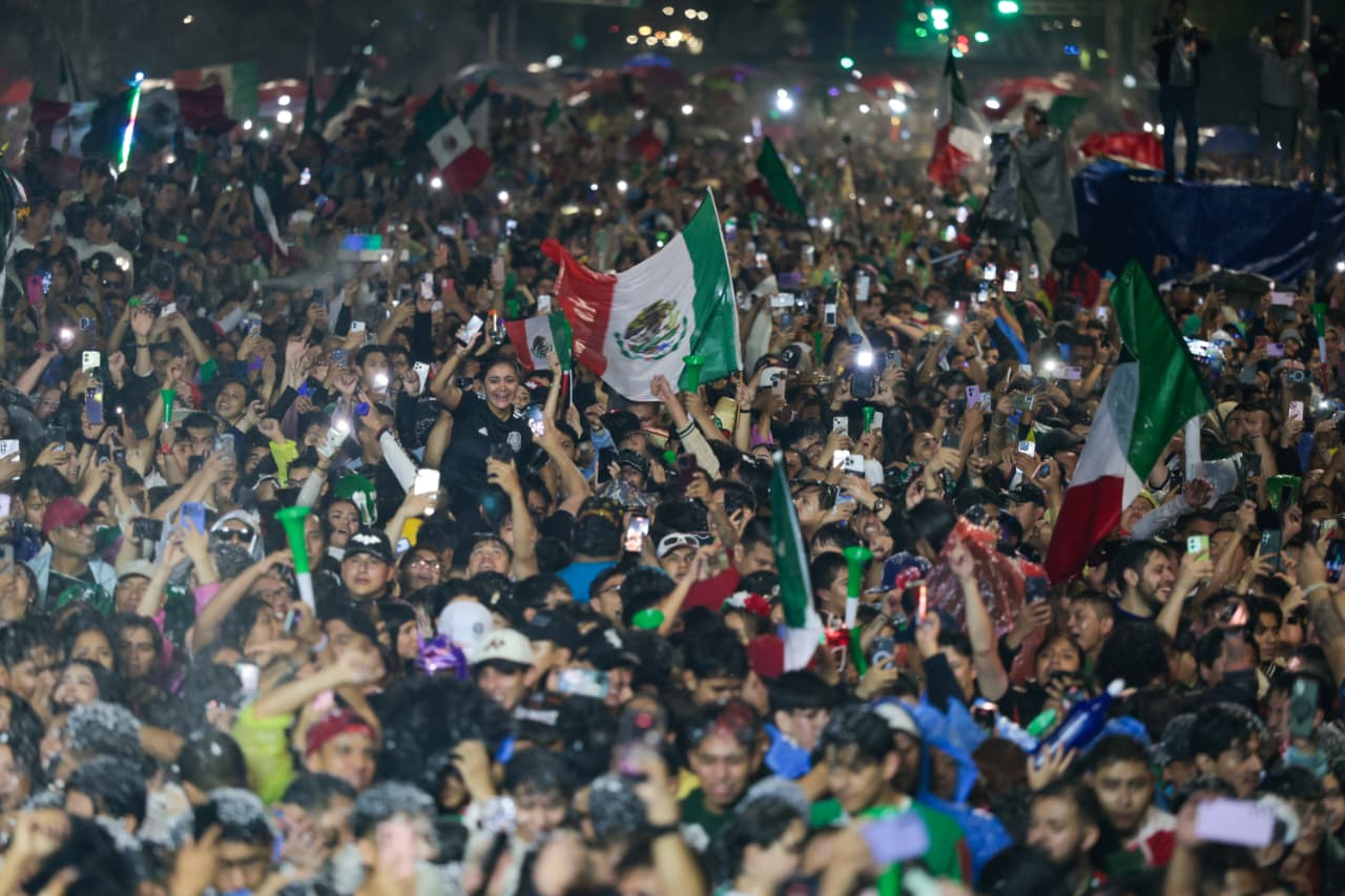 Una Toma Aérea Nocturna Que Captura Una Densa Multitud De Miles De Aficionados Al Fútbol Celebrando En La Avenida Paseo De La Reforma De La Ciudad De México. La Escena Es Un Mar De Color Y Emoción, Con Innumerables Personas Ondeando Banderas Mexicanas Verdes, Blancas Y Rojas. Muchas Personas En Primer Plano Están Levantando Sus Teléfonos Celulares Iluminados, Creando Un Efecto Brillante En Toda La Multitud. Las Personas Tienen Expresiones De Alegría Y Están Coreando. La Avenida Está Completamente Llena Hasta Donde Alcanza La Vista, Con Semáforos Verdes Y Luces De La Ciudad Borrosas Al Fondo, Subrayando La Escala Del Evento Masivo. La Imagen Captura La Euforia Colectiva Y La Unidad De Los Aficionados Mexicanos.