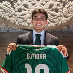 Retrato De Medio Cuerpo Del Futbolista Mexicano Gilberto Mora. El Joven Sonríe A La Cámara Vistiendo Un Traje Formal Negro, Camisa Blanca Y Corbata Verde Oscuro. Con Ambas Manos Sostiene Frente A Él La Parte Posterior De La Playera Verde De La Selección Mexicana, Donde Se Lee Claramente El Nombre Impreso &Amp;Quot;G. Mora&Amp;Quot; En Letras Blancas Sobre El Número 10. Al Fondo, Se Aprecia Una Imponente Pieza Arqueológica Tallada En Piedra Con Relieves Prehispánicos Y Una Pared De Bloques Texturizados En Tonos Marrón.