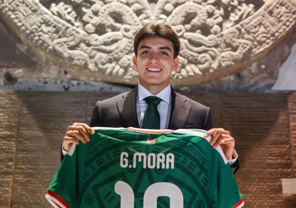 Gilberto mora: el orgullo de chiapas se convierte en el jugador más joven de la copa mundial 2026 la revista del sureste mx Retrato De Medio Cuerpo Del Futbolista Mexicano Gilberto Mora. El Joven Sonríe A La Cámara Vistiendo Un Traje Formal Negro, Camisa Blanca Y Corbata Verde Oscuro. Con Ambas Manos Sostiene Frente A Él La Parte Posterior De La Playera Verde De La Selección Mexicana, Donde Se Lee Claramente El Nombre Impreso &Amp;Quot;G. Mora&Amp;Quot; En Letras Blancas Sobre El Número 10. Al Fondo, Se Aprecia Una Imponente Pieza Arqueológica Tallada En Piedra Con Relieves Prehispánicos Y Una Pared De Bloques Texturizados En Tonos Marrón.