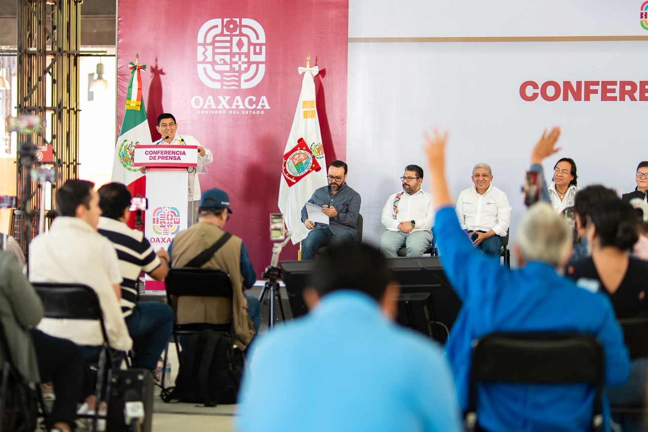 Fotografía En Plano General Que Muestra El Desarrollo De Una Rueda De Prensa En Un Auditorio. Al Fondo A La Izquierda, El Gobernador De Oaxaca, Salomón Jara Cruz, Se Encuentra De Pie Hablando Detrás De Un Podio Oficial Que Exhibe La Leyenda &Quot;Conferencia De Prensa&Quot;.