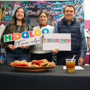 Fotografía De Plano Medio Que Muestra A Cinco Funcionarios Públicos Alineados Detrás De Una Mesa Negra Durante Una Rueda De Prensa Institucional. En Los Extremos, Un Hombre Y Una Mujer Visten Chalecos Institucionales De Color Guinda. Al Centro, Una Joven Con Chaqueta De Piel Negra Sostiene Un Letrero Colorido Que Dice &Amp;Quot;Hidalgo ¡Tiene Algo!&Amp;Quot;; A Su Lado, Otra Mujer Sostiene El Letrero Promocional Con El Nombre &Amp;Quot;Tianguistengo&Amp;Quot;, Y Junto A Ella, Un Hombre Con Chaleco Acolchado Azul Marino Sonríe A La Cámara. Sobre La Mesa Se Aprecian Vasos De Plástico Con La Bebida Tradicional Axocotl Y Un Platón De Barro Con Pan Artesanal Cielito Y Hojarascas. El Fondo Está Cubierto Por Una Gran Lona Publicitaria Con Fuegos Artificiales Y Los Detalles De La Feria Del 22 Al 26 De Julio.