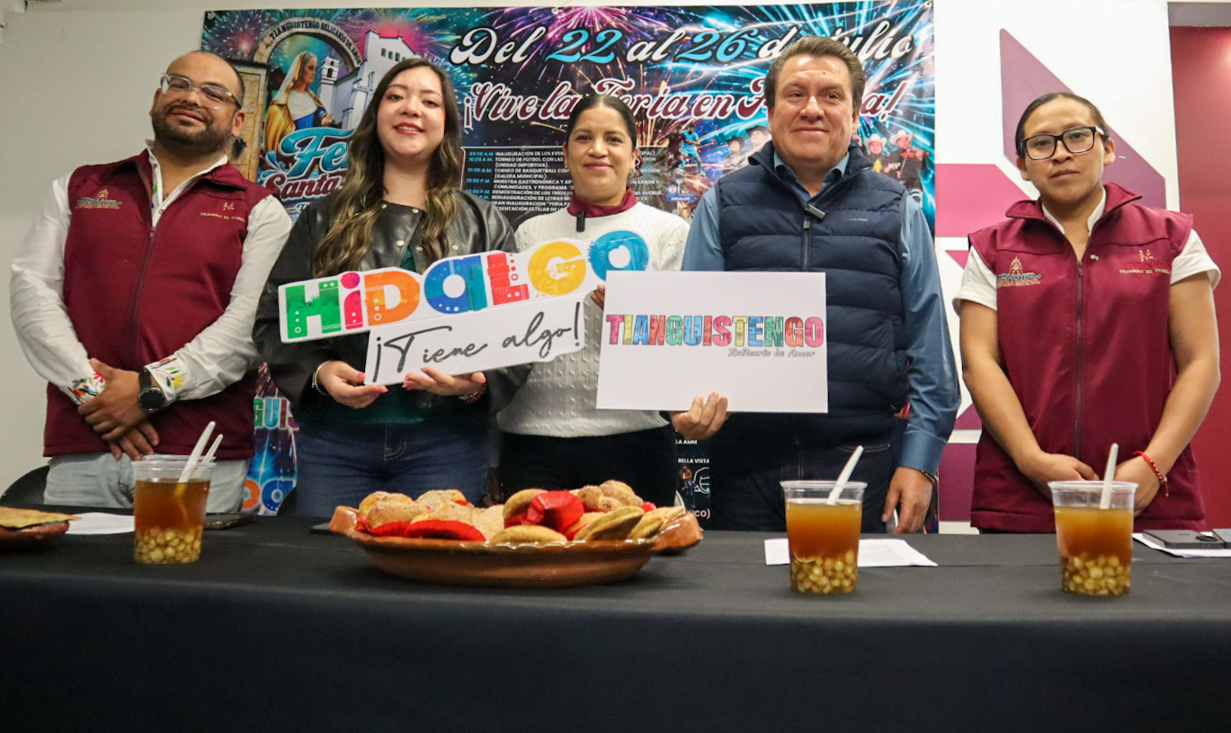 Fotografía De Plano Medio Que Muestra A Cinco Funcionarios Públicos Alineados Detrás De Una Mesa Negra Durante Una Rueda De Prensa Institucional. En Los Extremos, Un Hombre Y Una Mujer Visten Chalecos Institucionales De Color Guinda. Al Centro, Una Joven Con Chaqueta De Piel Negra Sostiene Un Letrero Colorido Que Dice &Amp;Quot;Hidalgo ¡Tiene Algo!&Amp;Quot;; A Su Lado, Otra Mujer Sostiene El Letrero Promocional Con El Nombre &Amp;Quot;Tianguistengo&Amp;Quot;, Y Junto A Ella, Un Hombre Con Chaleco Acolchado Azul Marino Sonríe A La Cámara. Sobre La Mesa Se Aprecian Vasos De Plástico Con La Bebida Tradicional Axocotl Y Un Platón De Barro Con Pan Artesanal Cielito Y Hojarascas. El Fondo Está Cubierto Por Una Gran Lona Publicitaria Con Fuegos Artificiales Y Los Detalles De La Feria Del 22 Al 26 De Julio.