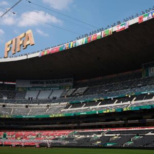 Una Vista De Ángulo Bajo Que Mira Hacia El Interior Del Estadio Ciudad De México (Anteriormente Estadio Azteca), Capturando Las Gradas Vacías Con Asientos Rojos Y Grises. Una Gran Estructura Colgante De Letras Doradas Que Deletrean &Amp;Quot;Fifa&Amp;Quot; Se Suspende De Cables En La Esquina Superior Izquierda Del Encuadre. Una Fila De Banderas Nacionales De Los Países Participantes Bordea La Viga Superior Del Techo, Y Un Letrero De &Amp;Quot;Fifa World Cup 2026&Amp;Quot; Es Visible En Una Sección De La Tribuna Media. El Campo De Juego Está Parcialmente Cubierto Por Equipos De Iluminación En Carretillas De Mano.