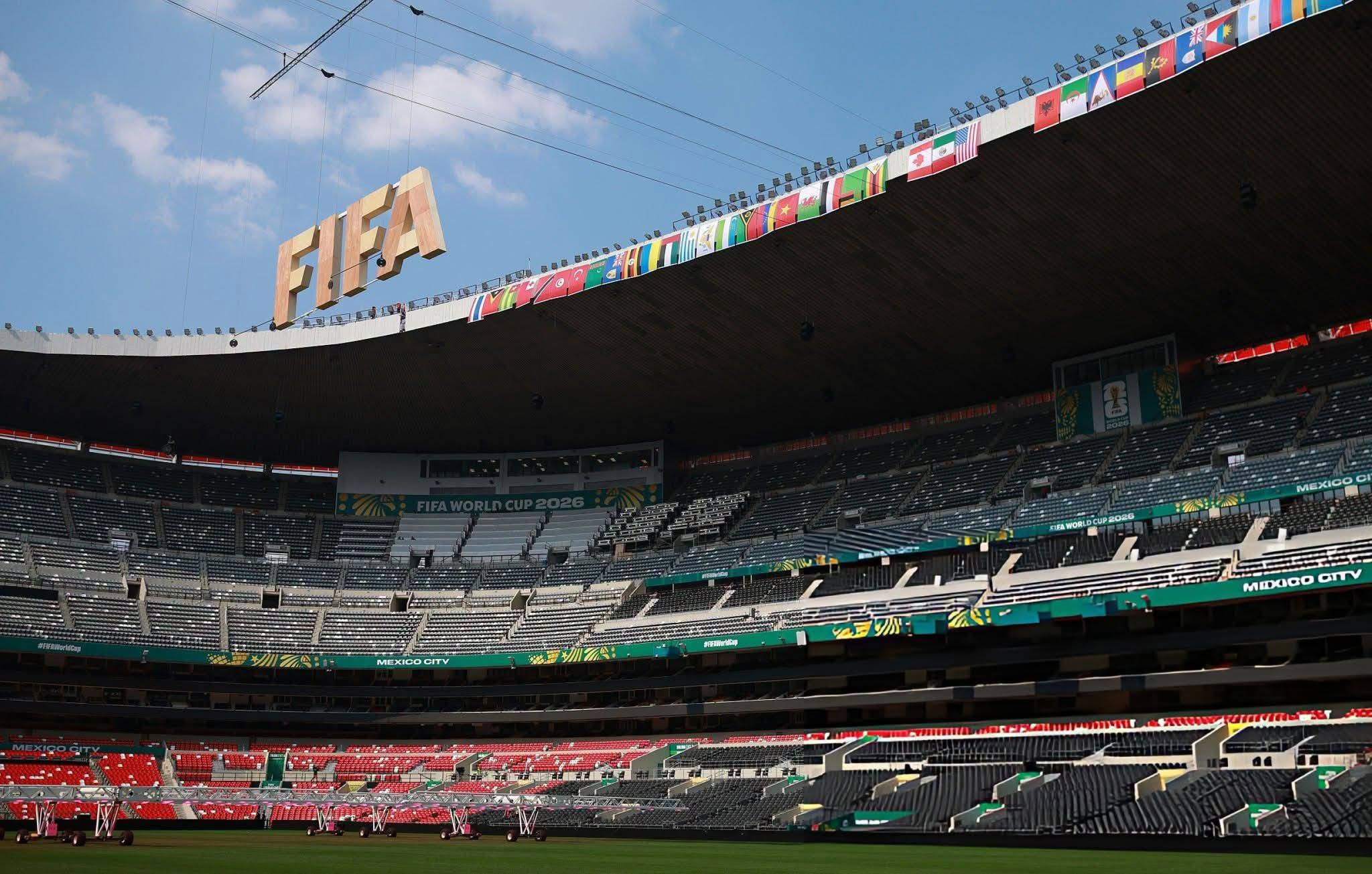 Una Vista De Ángulo Bajo Que Mira Hacia El Interior Del Estadio Ciudad De México (Anteriormente Estadio Azteca), Capturando Las Gradas Vacías Con Asientos Rojos Y Grises. Una Gran Estructura Colgante De Letras Doradas Que Deletrean &Amp;Quot;Fifa&Amp;Quot; Se Suspende De Cables En La Esquina Superior Izquierda Del Encuadre. Una Fila De Banderas Nacionales De Los Países Participantes Bordea La Viga Superior Del Techo, Y Un Letrero De &Amp;Quot;Fifa World Cup 2026&Amp;Quot; Es Visible En Una Sección De La Tribuna Media. El Campo De Juego Está Parcialmente Cubierto Por Equipos De Iluminación En Carretillas De Mano.