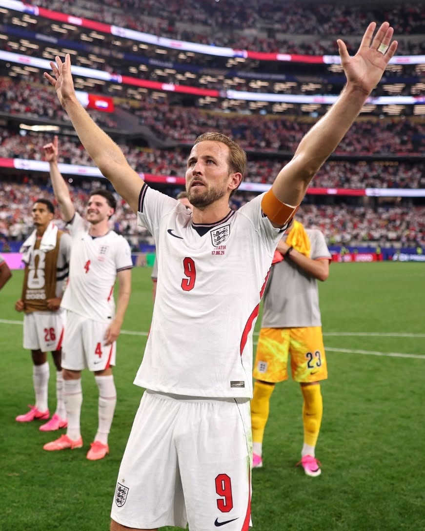 Plano Medio Del Delantero Y Capitán De La Selección De Inglaterra, Harry Kane, Celebrando En La Cancha Con Ambos Brazos Levantados Hacia El Cielo Tras Un Partido Del Mundial 2026. Viste El Uniforme Blanco Oficial De Los Tres Leones Con El Número 9 En Rojo En El Pecho Y El Short, Destacando Además La Cinta De Capitán En Su Brazo Izquierdo. Al Fondo Se Aprecian Las Tribunas De Un Estadio Lleno Y A Otros Compañeros De Equipo Festejando El Triunfo.