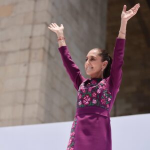 Hilos de identidad: el significado detrás del vestido oaxaqueño de la presidenta claudia sheinbaum la revista del sureste mx La Presidenta De México, Claudia Sheinbaum Pardo, Sonríe Con Los Brazos Levantados En Señal De Saludo Durante Un Evento Público. Viste Un Elegante Vestido De Manga Larga Color Fucsia Oscuro Con Detallados Bordados Florales Artesanales En Tonos Rosa, Morado Y Verde En El Pecho Y Los Costados. Al Fondo Se Observa Una Estructura De Piedra Clara Bajo La Luz Del Día.