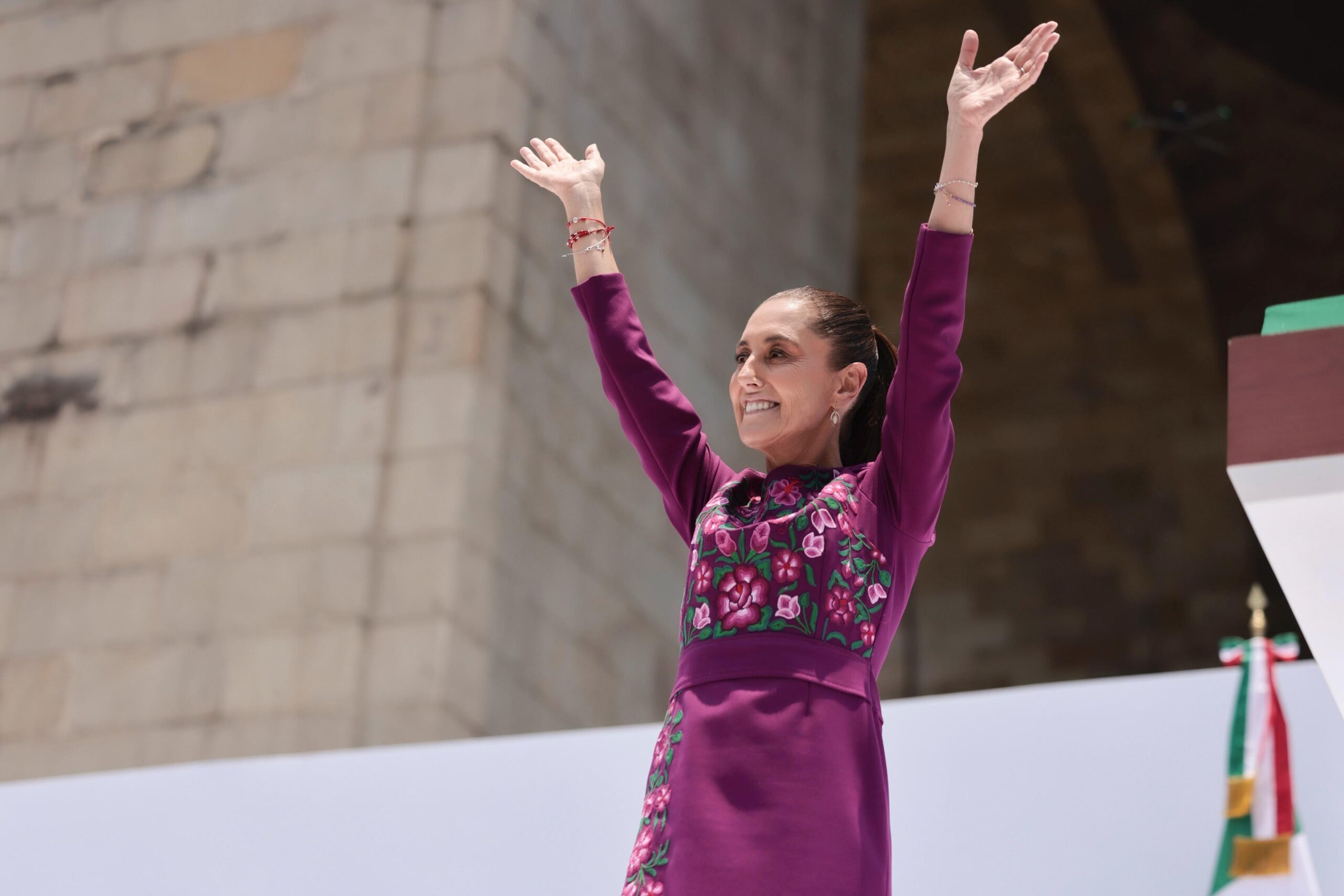 Hilos de identidad: el significado detrás del vestido oaxaqueño de la presidenta claudia sheinbaum la revista del sureste mx La Presidenta De México, Claudia Sheinbaum Pardo, Sonríe Con Los Brazos Levantados En Señal De Saludo Durante Un Evento Público. Viste Un Elegante Vestido De Manga Larga Color Fucsia Oscuro Con Detallados Bordados Florales Artesanales En Tonos Rosa, Morado Y Verde En El Pecho Y Los Costados. Al Fondo Se Observa Una Estructura De Piedra Clara Bajo La Luz Del Día.