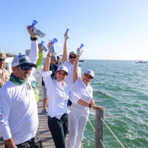 Fotografía En Plano Entero Que Muestra A Un Grupo De Personas, Encabezado Por La Presidenta Municipal Ana Paty Peralta, Celebrando El Inicio De Un Evento Sobre Un Muelle De Madera Junto Al Mar Turquesa. En El Centro De La Imagen, Ana Paty Peralta Sonríe Ampliamente Vistiendo Una Camisa Blanca De Manga Larga Y Pantalón Oscuro, Levantando Una Corneta De Aire Azul En Señal De Salida.