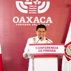 Fotografía En Plano Medio Del Gobernador De Oaxaca, Salomón Jara Cruz, Sonriendo Detrás De Un Podio Blanco Con Un Letrero En Letras Rojas Que Dice &Amp;Quot;Conferencia De Prensa&Amp;Quot;. Viste Una Guayabera Blanca Tradicional Con Sutiles Bordados Florales Amarillos Y Verdes Cerca Del Cuello. Al Fondo, Se Aprecia Una Pared De Color Guinda Con El Logotipo Institucional En Letras Blancas Que Dice &Amp;Quot;Oaxaca Gobierno Del Estado&Amp;Quot;.