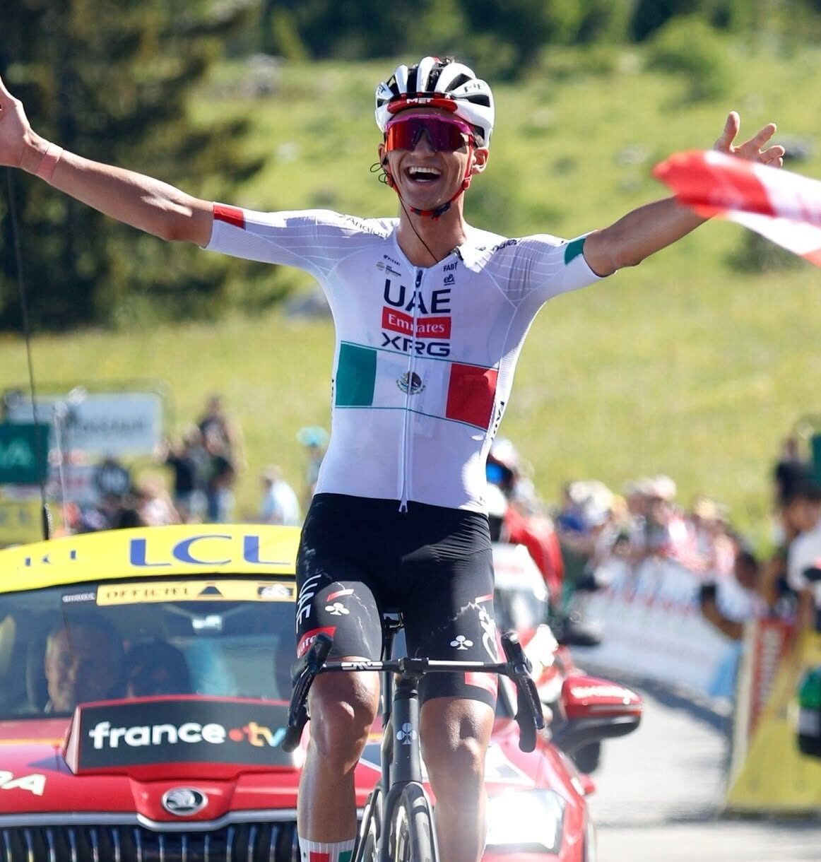 Fotografía En Plano Medio Y Ángulo Contrapicado Del Ciclista Mexicano Isaac Del Toro Celebrando Al Cruzar La Línea De Meta En Una Competencia De Ruta. El Atleta Aparece Montado En Su Bicicleta De Carreras Negra, Con Ambos Brazos Levantados, Las Manos Abiertas Y Una Expresión De Gran Felicidad Y Euforia.
