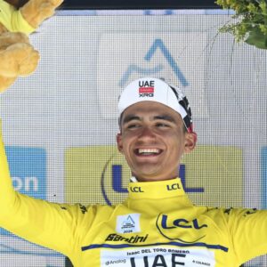 Fotografía En Plano Medio Del Ciclista Isaac Del Toro En El Podio De Premiación Tras Ganar Una Competencia. El Joven Deportista Sonríe Ampliamente Mientras Levanta Ambos Brazos En Señal De Victoria; En Su Mano Derecha Sostiene Un Peluche Del León De Lcl Y En Su Mano Izquierda Un Ramo De Flores Amarillas.