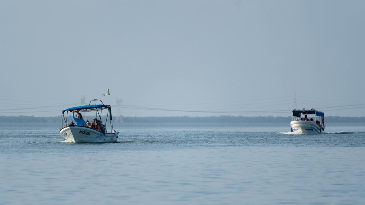 Una Vista Aérea De La Isla Lobos En Veracruz, Con Aguas Turquesas Y Arrecifes De Coral Visibles Bajo La Superficie. Dos Botes De Excursión Y Un Bote Más Pequeño Se Acercan A La Isla, Que Cuenta Con Un Faro Blanco Y Un Bosque De Palmeras.