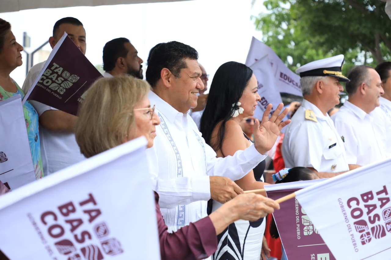 Fotografía En Plano Medio Lateral De Un Grupo De Autoridades Civiles Y Militares Durante Un Evento Público Al Aire Libre Bajo Una Lona Blanca. En El Centro De La Toma Destaca El Gobernador De Tabasco, Javier May Rodríguez, Vistiendo Una Guayabera Blanca Tradicional Y Sonriendo Mientras Sostiene Un Banderín Oficial.