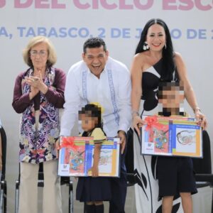 Fotografía En Plano Medio Largo Y Toma Frontal De Un Evento Institucional. Al Centro, El Gobernador De Tabasco, Javier May Rodríguez, Viste Guayabera Blanca Y Sonríe Con Entusiasmo Mientras Ayuda A Una Niña De Uniforme Escolar A Sostener Un Paquete Grande De Libros Envuelto En Celofán Con Un Moño Rojo.