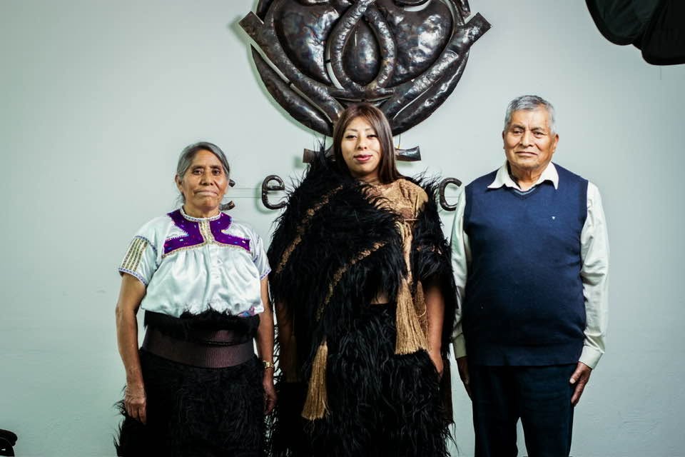 Fotografía De Susi Bentzulul En El Centro, Vistiendo Indumentaria Tradicional De San Juan Chamula Con Un Chuj Negro De Lana Y Detalles Dorados, Flanqueada Por Sus Padres. Al Fondo, Se Aprecia El Logotipo Metálico Del Cesmeca.