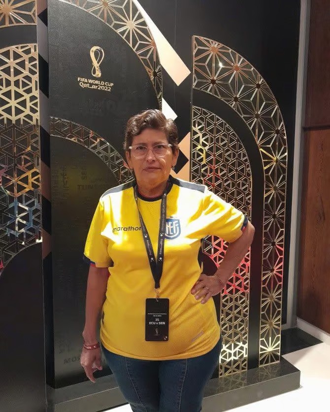 El boleto dorado del futbol: la mujer que lleva 28 años viajando gratis a todos los mundiales la revista del sureste mx Fotografía De Plano Medio De María Del Jesús Pinargote Posando De Pie Frente A Un Fondo Decorativo Oficial De La Copa Mundial De La Fifa Catar 2022. Viste La Playera Amarilla De La Selección De Ecuador De La Marca Marathon Con El Escudo De La Fef Y Pantalones De Mezclilla Azules. Tiene El Cabello Corto, Usa Lentes Transparentes Y Tiene La Mano Izquierda Apoyada En La Cadera. Al Cuello Lleva Colgada Una Acreditación Oficial De Color Negro Con La Leyenda &Quot;Ecu V Sen&Quot;. El Fondo Es Oscuro Con Estructuras Arqueadas Que Muestran Intrincados Patrones Geométricos Y Calados En Color Dorado Junto Al Logotipo Dorado De Qatar 2022.