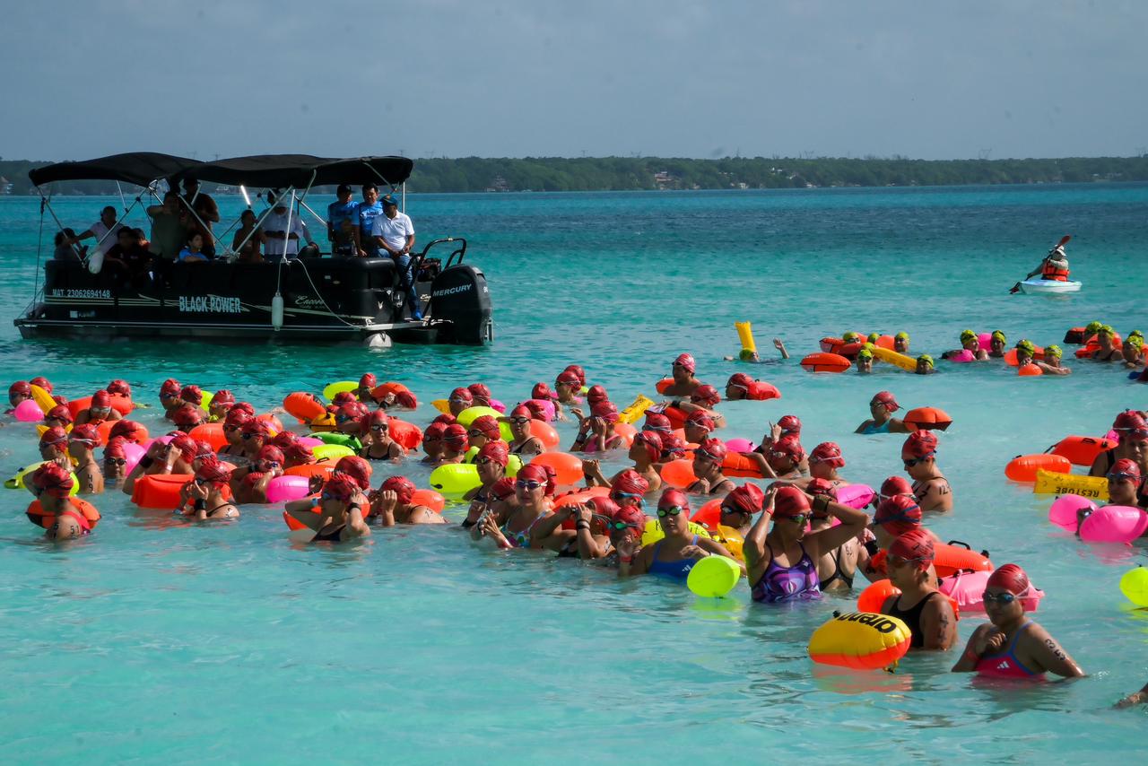 Mara lezama encabeza maratón de aguas abiertas en bacalar la revista del sureste mx Una Vista Aérea De Un Gran Grupo De Nadadores Con Gorros De Natación De Colores Brillantes, Principalmente Naranjas Y Rosas, Y Boyas De Seguridad Amarillas Flotando A Su Alrededor, Preparándose Para Una Carrera En Aguas Abiertas En Un Cuerpo De Agua Turquesa Brillante, Que Es La Laguna De Bacalar. Los Nadadores Están Densamente Agrupados En Primer Plano Y Medio Plano, Mirando Hacia Adelante.