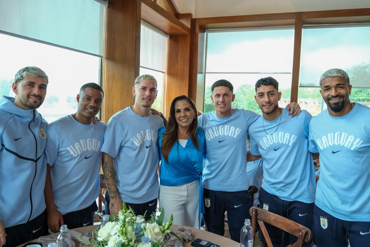 Fotografía De Plano Medio En Interiores Que Muestra A La Gobernadora Mara Lezama En El Centro De Un Segundo Grupo De Seis Futbolistas De La Selección Nacional De Uruguay. La Gobernadora Sonríe A La Cámara Vistiendo Su Conjunto De Blusa Azul Rey Y Pantalón Blanco, Con El Brazo Izquierdo Alrededor Del Hombro Del Mediocampista Federico Valverde, Quien Viste La Playera Azul De Entrenamiento De La Delegación Uruguaya.