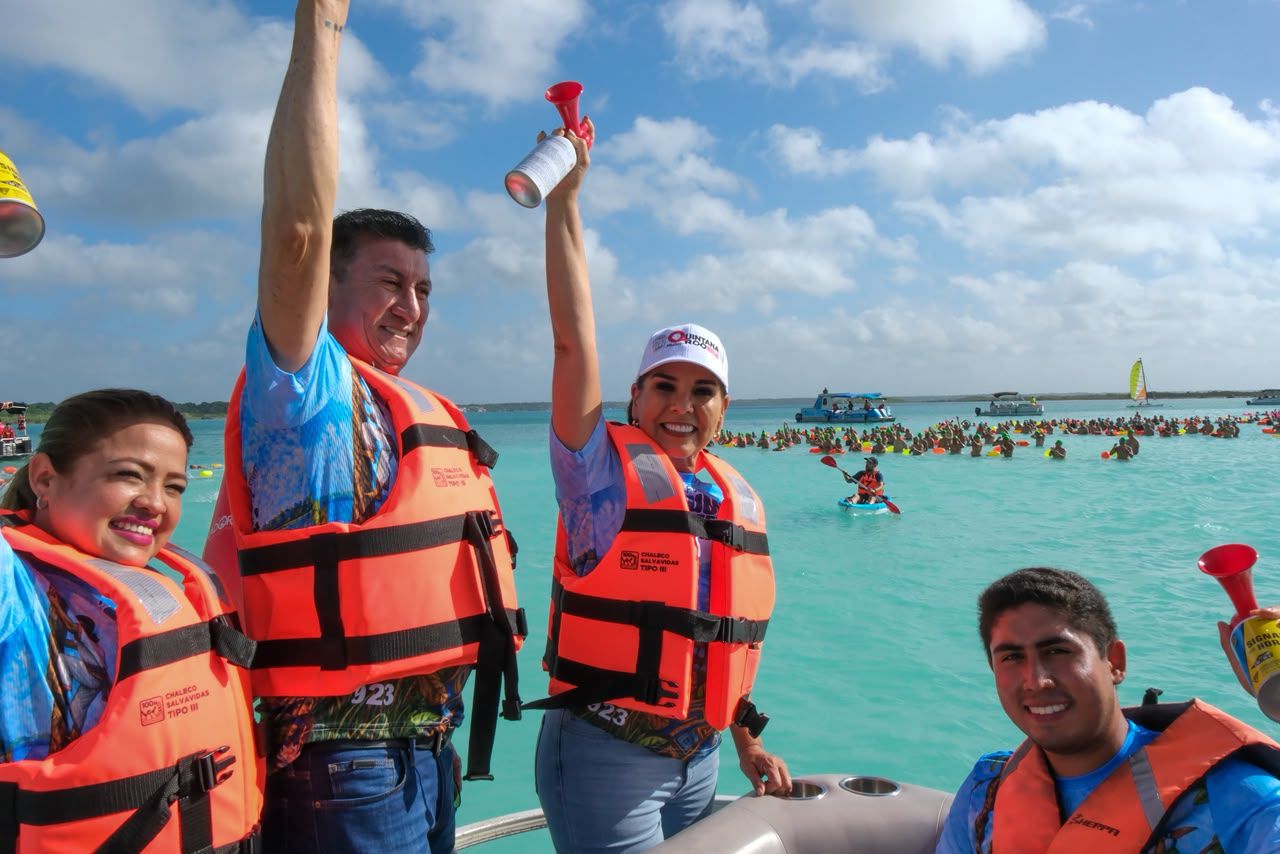 La Gobernadora De Quintana Roo, Mara Lezama Espinosa, Sonríe En El Centro Mientras Levanta Una Bocina De Aire Roja Para Dar El Banderazo De Salida. Viste Jeans, Una Gorra Blanca Con El Logotipo Del Estado Y Un Chaleco Salvavidas Naranja Brillante Tipo Iii Sobre Una Camiseta Azul Con Estampados Tropicales.