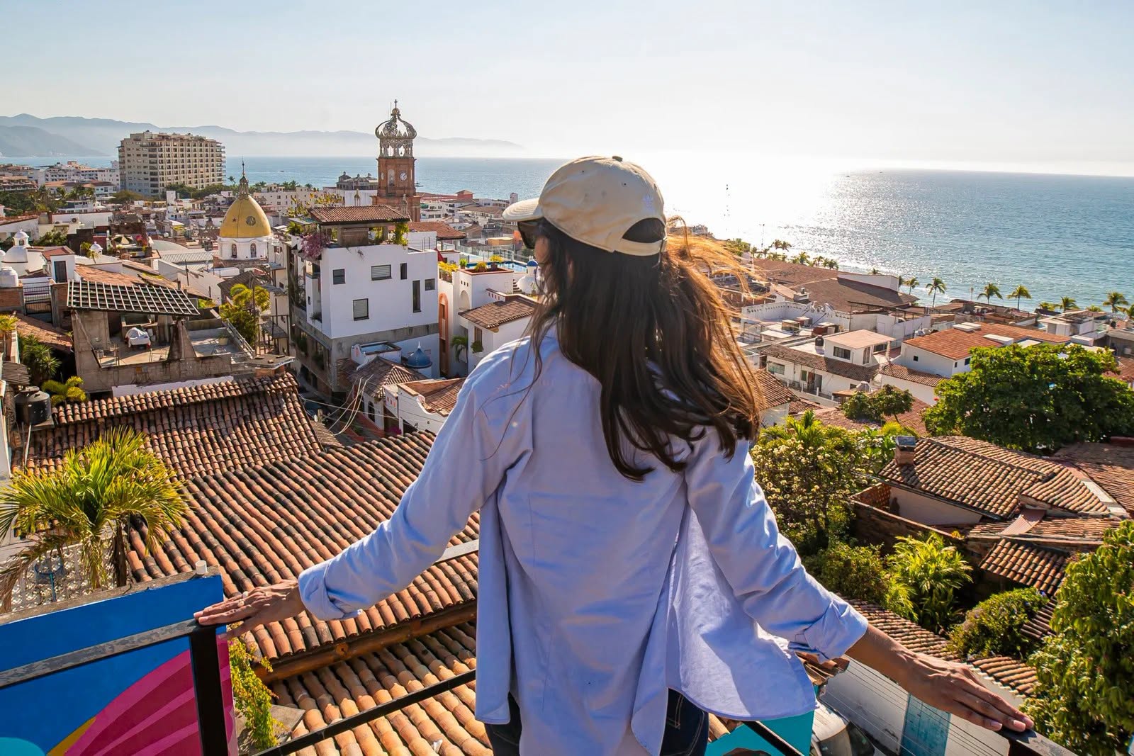 El orgullo de méxico: por qué jalisco conquista al mundo y es nombrado "mejor destino internacional 2026" por condé nast traveler la revista del sureste mx Una Vista Panorámica En Un Día Soleado De Una Ciudad Costera Con Tejados De Terracota Y Una Costa Oceánica A La Derecha, Vista Por Una Joven Viajera De Espaldas Que Lleva Una Gorra De Béisbol De Color Claro Y Una Camisa Azul Desabrochada Sobre Otra Camiseta. Ella Está De Pie Detrás De Una Barandilla Metálica, Con Las Manos Apoyadas En Ella, Mirando Hacia El Centro Histórico De La Ciudad, Que Cuenta Con Una Distintiva Iglesia Con Cúpula Dorada Y Una Torre De Reloj Más Alta.