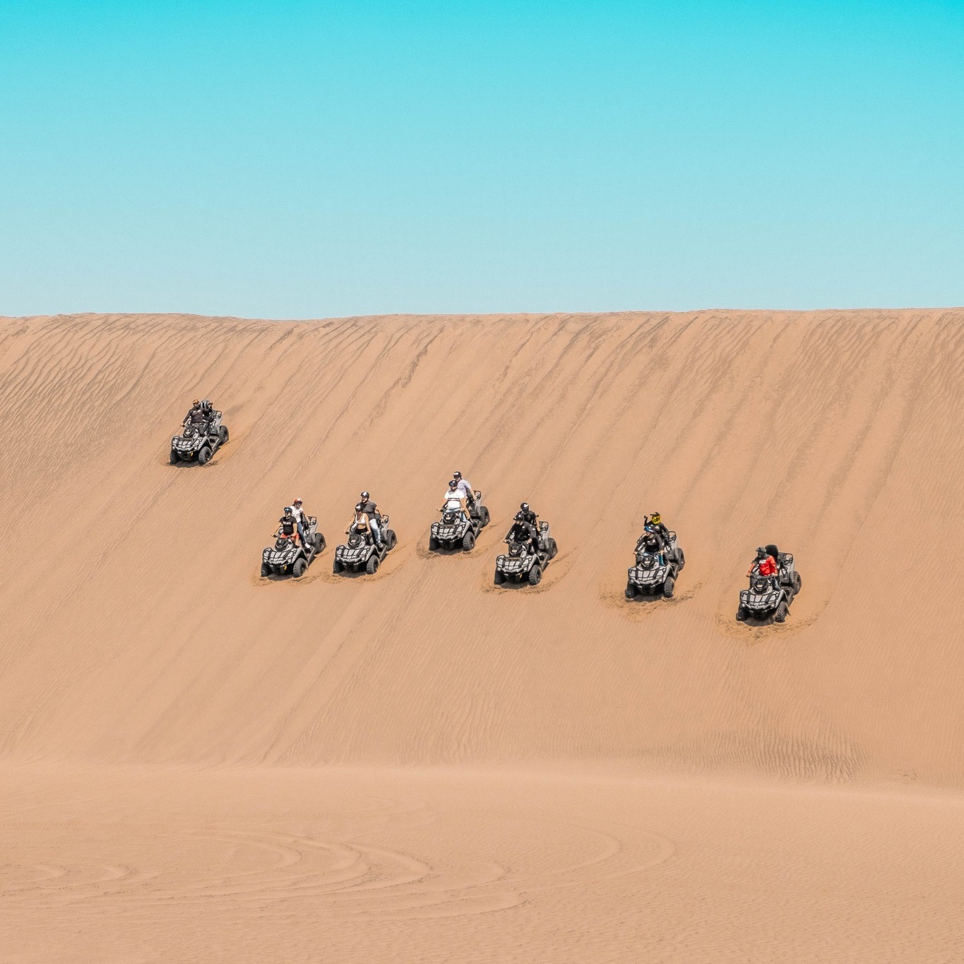 Una Toma De Arriba Hacia Abajo De Un Grupo De Siete Vehículos Todo Terreno Circulando En Formación Por Una Empinada Duna De Arena En El Desierto. Los Atvs Y Sus Conductores, Todos Con Equipo De Seguridad, Son De Varios Colores Y Tamaños. El Cielo Azul Claro Y Soleado Se Extiende Por Encima, Y Las Ondulaciones En La Arena Crean Un Patrón Texturizado.