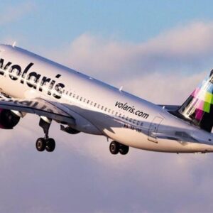 Fotografía En Plano Medio De Un Avión Comercial De La Aerolínea Volaris En Pleno Despegue, Elevándose Diagonalmente Hacia La Izquierda Sobre Un Fondo De Cielo Azul Con Nubes Suaves De Tonos Rosados. El Avión Cuenta Con Fuselaje Blanco Donde Se Aprecia La Marca &Amp;Quot;Volaris&Amp;Quot; En Letras Negras, Motores De Color Fucsia Brillante Bajo Las Alas Y Una Cola De Color Negro Con Un Diseño Geométrico De Cuadros En Tonos Fucsia, Verde Y Azul. Esta Imagen Ilustra El Crecimiento De La Conectividad Aérea En México Y La Apertura De Nuevas Rutas Comerciales.