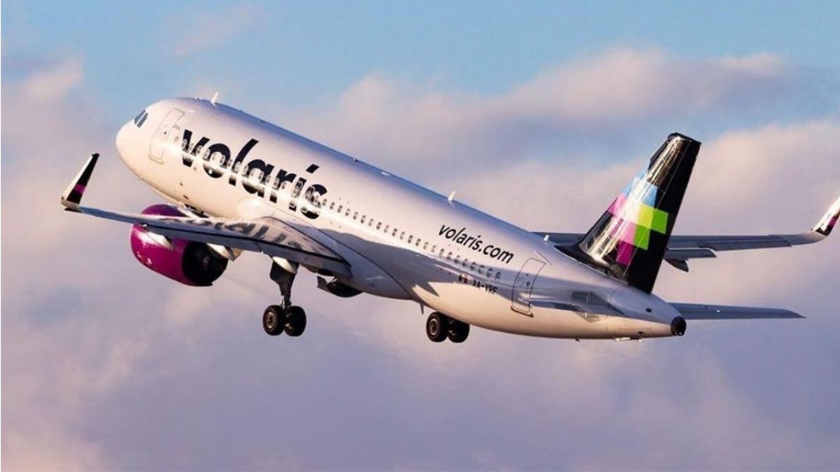 Fotografía En Plano Medio De Un Avión Comercial De La Aerolínea Volaris En Pleno Despegue, Elevándose Diagonalmente Hacia La Izquierda Sobre Un Fondo De Cielo Azul Con Nubes Suaves De Tonos Rosados. El Avión Cuenta Con Fuselaje Blanco Donde Se Aprecia La Marca &Quot;Volaris&Quot; En Letras Negras, Motores De Color Fucsia Brillante Bajo Las Alas Y Una Cola De Color Negro Con Un Diseño Geométrico De Cuadros En Tonos Fucsia, Verde Y Azul. Esta Imagen Ilustra El Crecimiento De La Conectividad Aérea En México Y La Apertura De Nuevas Rutas Comerciales.
