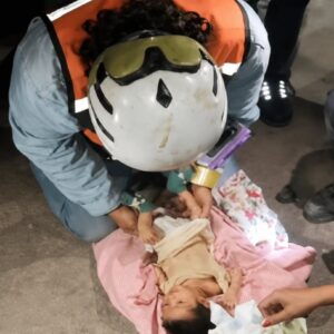 Fotografía En Picada Que Muestra A Un Brigadista De Rescate Agachado Sobre El Suelo, Atendiendo A Un Bebé Recién Nacido Recostado Sobre Una Manta Rosa. El Rescatista Usa Un Casco Blanco De Protección Con Gogles Amarillos Integrados, Una Chamarra Azul Claro Y Un Chaleco Reflejante Naranja. Con Sus Manos Acomoda Con Delicadeza La Ropa Del Lactante, Quien Se Encuentra Bocarriba Con El Rostro Visible. Alrededor, Se Observan Las Piernas Y Manos De Otras Personas Del Equipo De Emergencias Brindando Asistencia En Un Entorno Nocturno Con Suelo De Concreto.
