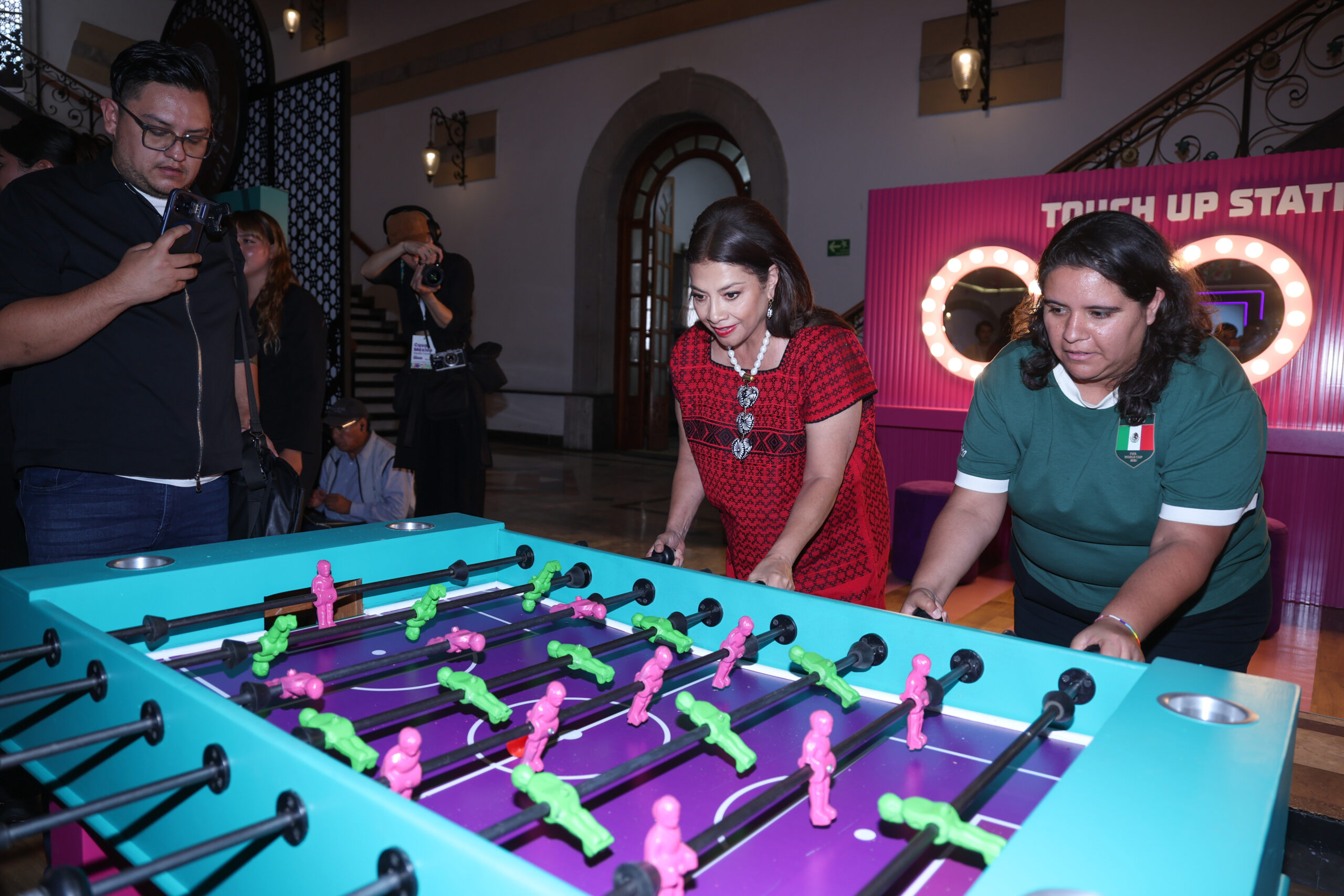 Mundial 2026: clara brugada inaugura centro de medios y presenta operativos en cdmx la revista del sureste mx Mundial 2026 Clara Brugada Inaugura Centro De Medios Y Presenta Operativos En Cdmx , La Jefa De Gobierno De La Ciudad De México, Clara Brugada Molina (En El Centro, Con Blusa Bordada Roja), Está Sonriendo Mientras Juega Una Partida De Futbolito. Está De Pie Junto A Otra Mujer Que Lleva Una Camiseta Verde De La Selección Nacional De México. El Futbolito Tiene Un Diseño Personalizado Con Bordes Turquesa Y Jugadores Pintados En Verde Neón Y Rosa. Al Fondo A La Izquierda, Un Hombre Con Gafas Sostiene Un Teléfono Inteligente, Y Otra Persona Con Una Cámara Profesional Está En La Parte Trasera. A La Derecha, Detrás Del Futbolito, Hay Una Estación Rosa De &Quot;Retoque De Imagen&Quot; Con Espejos Iluminados. La Escena Transcurre En Un Interior Con Arquitectura Histórica De La Ciudad De México, Visible En Las Escaleras Y Los Arcos De Piedra Al Fondo.