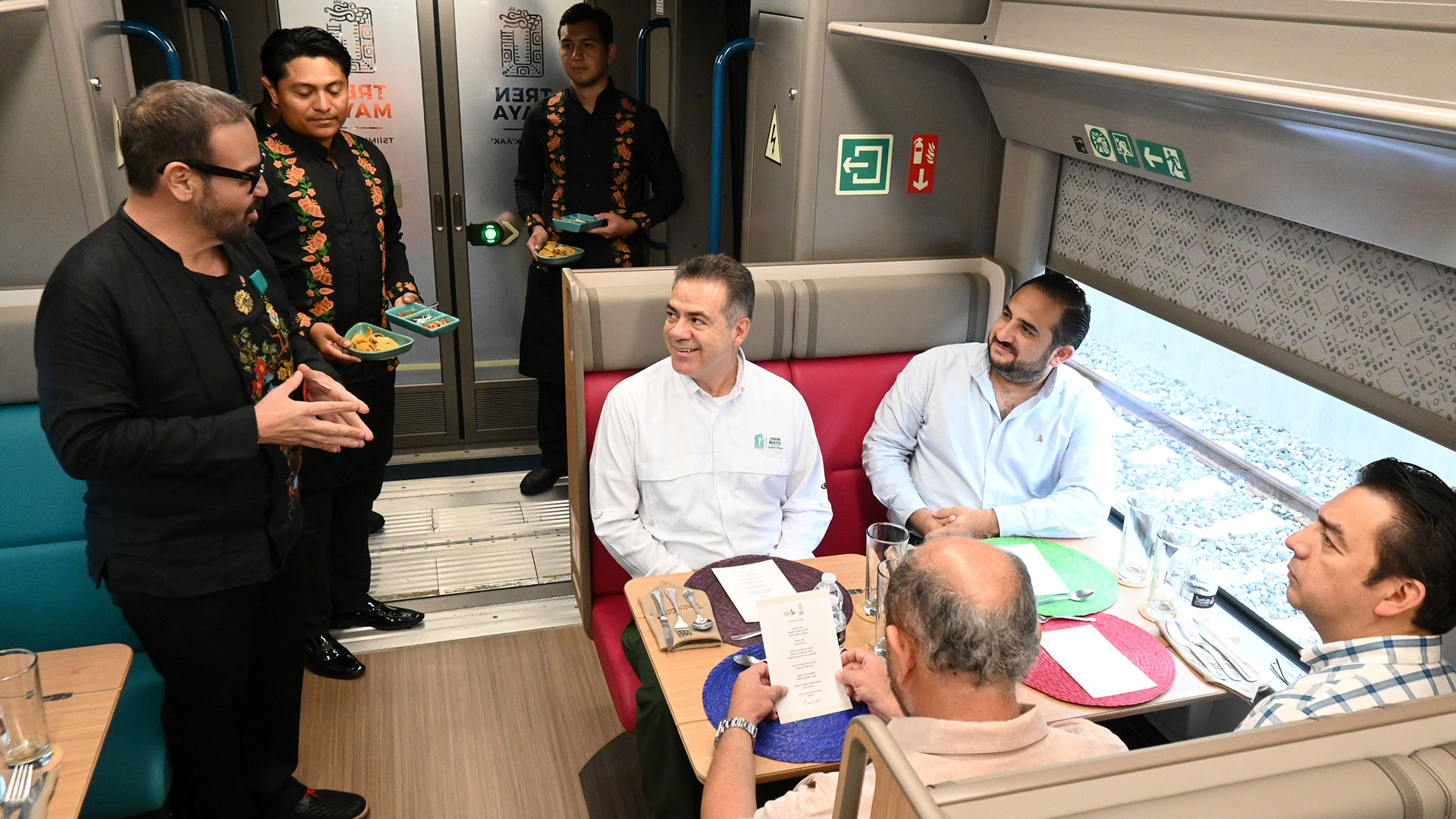 Fotografía En Plano General De Toma Picada En El Interior De Un Vagón Restaurante Del Tren Maya. A La Izquierda, Un Hombre De Lentes Y Camisa Negra Con Bordados Florales De Colores Habla De Pie Gesticulando Con Las Manos. Detrás De Él, Dos Meseros Con Camisas Negras Ornamentadas Con Detalles Tradicionales Llevan Platos Pequeños.