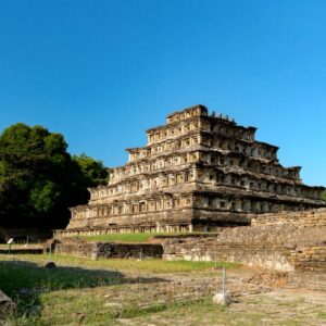 Fotografía A Cielo Abierto Que Muestra En Un Plano General La Pirámide De Los Nichos En La Zona Arqueológica De El Tajín. La Estructura Piramidal Está Compuesta Por Múltiples Niveles Escalonados Construidos En Piedra Labrada, Destacando Las Filas Simétricas De Nichos Cuadrangulares En Cada Una De Sus Caras. A La Izquierda De La Pirámide Se Observa Una Densa Vegetación De Árboles Verdes, Mientras Que En Primer Plano Se Aprecian Restos De Muros Y Estructuras Arqueológicas Menores Rodeadas De Pasto Corto. El Cielo Es Completamente Despejado Y De Un Color Azul Intenso, Iluminando La Escena Con Luz Solar Directa.